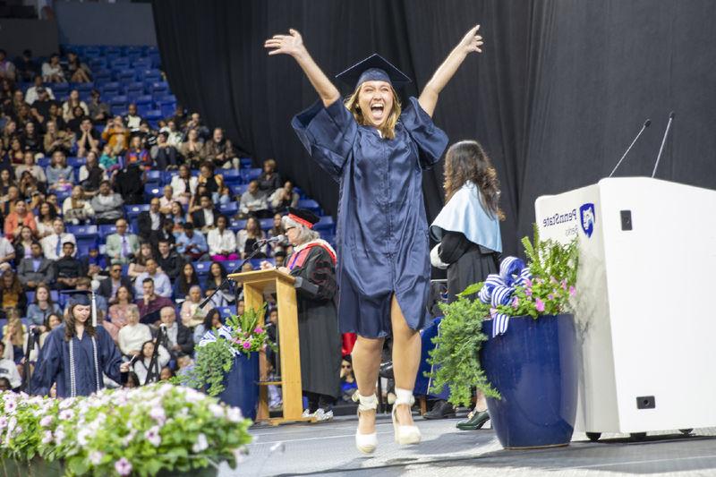 Student wearing cap and gown jumps for joy crossing the stage at commencement