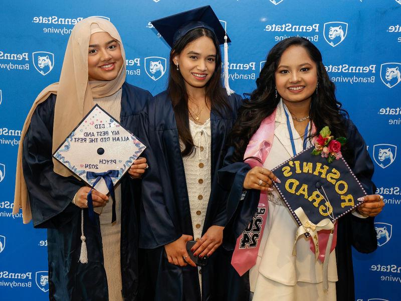 Three female students pose for a photo wearing their commencement gowns.