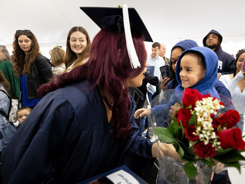 Woman in commencement gown bends down to accept a bouquet of flowers from a young boy.