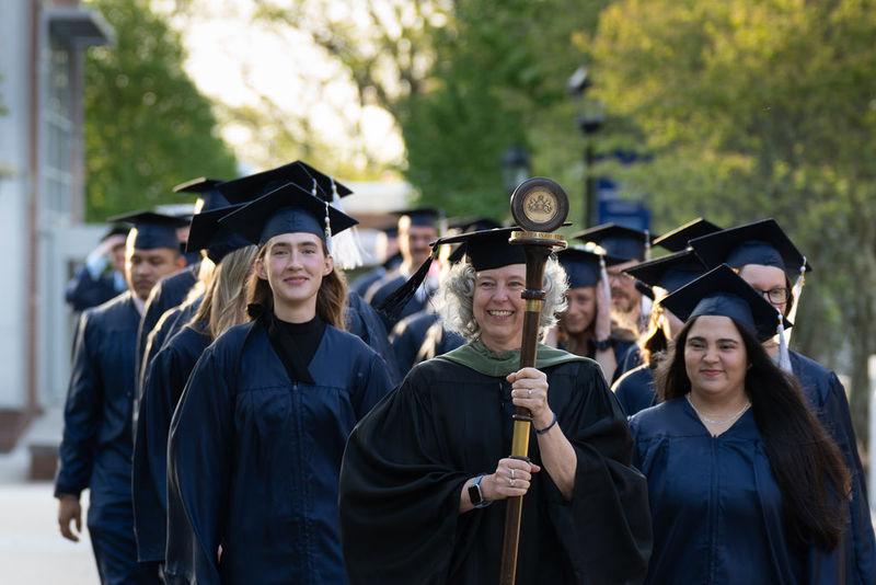 Graduates in caps and gowns in two rows walking down sidewalk led by faculty member in black robe carrying a staff and pendant.